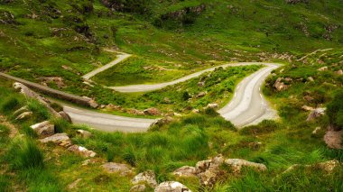 Dar bir yol, yemyeşil bir tepe boyunca keskin bir şekilde esiyor. Kayalar ve uzun otlar yol boyunca büyürler. Dunloe Geçidi güzel manzarasını gösteriyor..