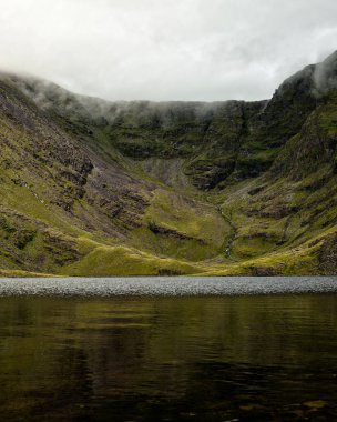 Bulutlar, dramatik dağların zirvelerini okşar. Yansımaları aşağıdaki durgun suda dans eder..