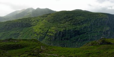 Yeşil tepeler yükselir ve dağlık bir bölgede sarp kayalıklar yaratır. Güneş ışığı bulutlu gökyüzüne nüfuz eder ve dağ yamaçlarındaki bazı ıslak alanları aydınlatır..