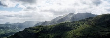 Dalgalanan yeşil yamaçları ve dramatik bulutlarla dolu bir gökyüzünün altındaki Dunloe Geçidi 'nin heybetli dağlarını gösteren manzaralı bir manzara. Yağmur kuyusu uzaklıktan görülebilir..