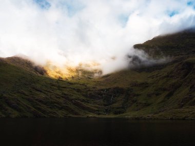 İrlanda 'nın kırsal kesimindeki Lough Callee yakınlarında kısmen beyaz sisle kaplanmış bir dağa yükselen yuvarlanan, çimlerle kaplı tepeler arasında durgun bir su kütlesi yer alır..