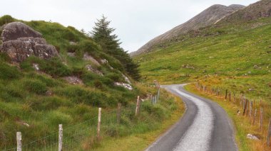 İrlanda 'nın Kerry ilçesindeki Dunloe' nin manzaralı geçidinde bulutlu bir gökyüzünün altında virajlı bir yol kıvrılır..