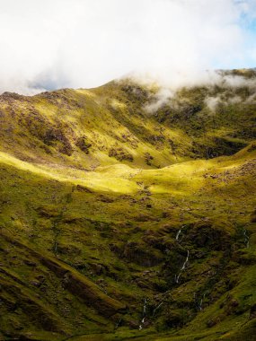 Bulutlu bir günde İrlanda 'nın Kerry ilçesindeki Carrauntoohil dağ yolunda dramatik yeşil manzara ve seyrek şelaleler görülüyor..