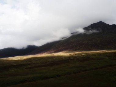 Bulutlu bir gökyüzünün altındaki Carrauntoohill Dağı 'nın güzel manzarası. Dağın eteğindeki yeşil tarlalarda ışık parlıyor. Bulutlar engebeli, kayalık arazinin zirvesini kaplar..