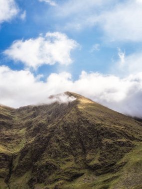 Görkemli bir dağ, tüylü beyaz bulutlarla benekli parlak mavi bir gökyüzüne doğru yükseliyor. Zirve, kısmen sis tarafından gizleniyor ve İrlanda 'daki Carrauntoohill' e gizemli bir kalite katıyor..