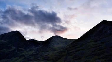 Carrauntoohill 'in sivri tepeleri, İrlanda' nın Kerry ilçesinde karanlık, dramatik bulutlar ve alacakaranlıkta kaybolan ışıklarla dolu bir gökyüzünün altında siluette görülür..