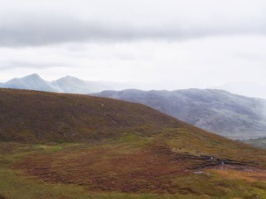 Tepeler kısmen kahverengi ve yeşil bitki örtüsüyle kaplıdır. Dağlar bu İrlanda manzarasında bulutlu ve bulutlu bir gökyüzünün altında görülebilir..