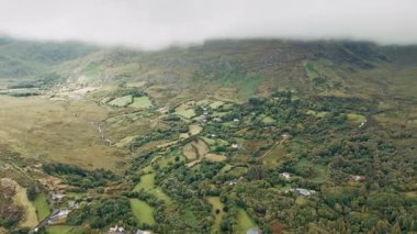 Yukarıdan, İrlanda 'nın Kerry ilçesinin çarpıcı manzarasını izleyin. Dağlar, ağaçlar, tarlalar ve küçük bir köy görülebilir durumda. Hava bulutlu ve bulutlu görünüyor..