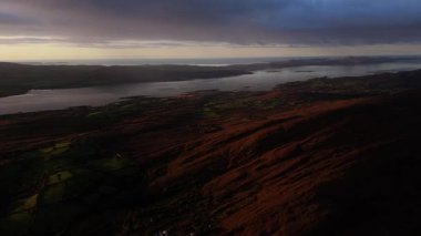Yükselmiş bir perspektif çarpıcı bir kıyı manzarasını yakalar. Dağlar, bulutlu bir günbatımı gökyüzünün altında geniş bir su kütlesine kadar uzanır..