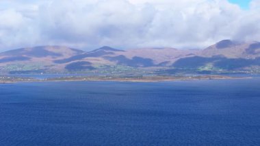 Geniş, pitoresk bir gündüz manzarası, mavi Atlantik Okyanusu 'nun İrlanda' nın Kerry ilçesinde kıyılarla buluşmasını, dağları ve bulutlu bir gökyüzünün altında yuvarlanan yeşil tepeleri gösteriyor..