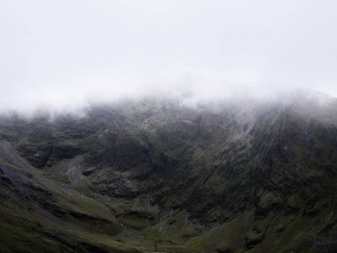 Bir dağ yüzeyi, alçak sis veya sis tarafından kısmen gizlenir. Yeşil yamaçlar ve kayalık kaya oluşumları beyaz gökyüzünün altında dramatik bir doku oluşturur..
