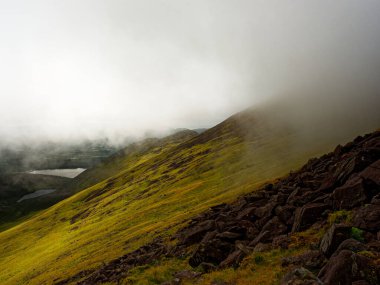 Yeşil yamaçlar İrlanda 'daki Coomloughra At Nalı' nda kayalık araziyle karşılaşıyor. Alçak bulutlar ve sis dağların tepelerini karartmış, aşağıdaki göllerle birlikte karamsar bir atmosfer yaratmış..
