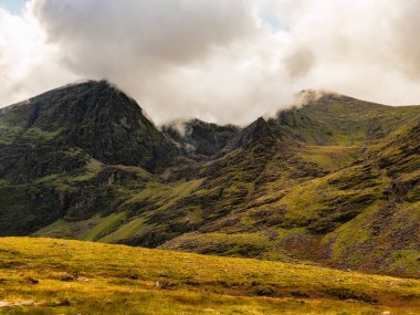 İrlanda 'nın Kerry ilçesindeki Carrauntoohil ilçesinde yemyeşil yamaçlar ve dağlık arazi görülüyor. Bulutlu gökyüzü İrlanda 'nın en yüksek zirvesinde hayranlık uyandıran bir manzara yaratır..