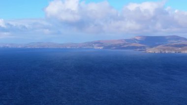 Güneşli bir öğleden sonra İrlanda 'nın yanındaki engin, derin mavi okyanus manzarası. Parçalı bulutlu bir gökyüzünün altında yuvarlanan tepeler görülebilir..