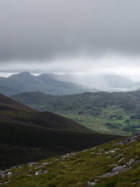 Bu manzara, Coomloughra at nalı yürüyüş yolu boyunca uzanan engebeli İrlanda manzarasını gösteriyor. Dağlar bulutlu bir gökyüzünün altında solup gidiyor, aşağıda da yeşil vadiler var..
