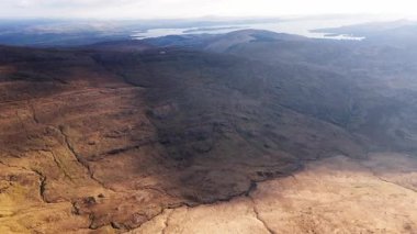 İrlanda, County Cork 'taki rahibin sıçrayışı yakınlarındaki engebeli araziyi araştırın. Çorak arazileri ve bulutlu bir gökyüzünün altındaki uzak gölleri gün boyunca yüksek bir perspektiften gözlemleyin..