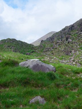 Dunloe Geçidi 'ndeki kayalık, yeşil manzaranın manzarası. Yuvarlanan yamaçlar ve büyük kayalar İrlanda kırsalında parçalı bulutlu bir gökyüzünün altında..