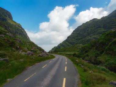 Kıvrımlı bir yol, gür yeşil bir vadiden geçer, kabarık beyaz bulutlarla benekli, canlı mavi bir gökyüzünün altında, heybetli dağların arasına yerleşir. Sahne bir macera ve sükunet hissi uyandırıyor..