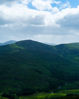 Sakin bir manzara, İrlanda 'nın Dunloe Geçidi' nde parçalı bulutlu bir gökyüzünün altında yuvarlanan yeşil tepeleri ve vadileri gösteriyor. Dağ ve vadi huzurlu ve manzaralı bir manzara yaratır..
