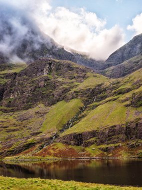 Sakin göl İrlanda 'daki Carrauntoohil dağlarını yansıtır. Sis ve bulutlar zirveyi gizliyor, bulutlu bir günde atmosferik bir manzara yaratıyor..