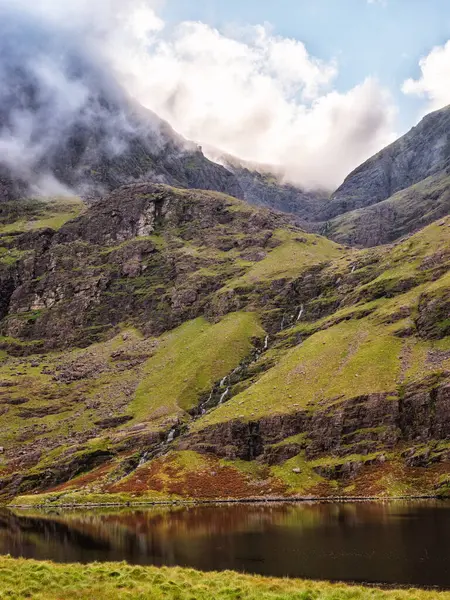 Sakin göl İrlanda 'daki Carrauntoohil dağlarını yansıtır. Sis ve bulutlar zirveyi gizliyor, bulutlu bir günde atmosferik bir manzara yaratıyor..