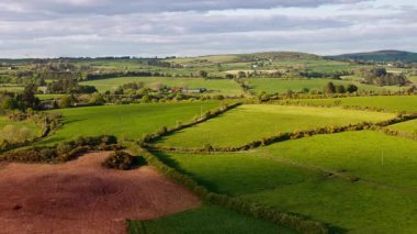 Hava perspektifi, İrlanda 'nın West Cork kırsalında, parçalı bulutlu bir gökyüzünün altında, çitlerle ayrılmış yeşil otlakları ve ekili alanları gösterir..