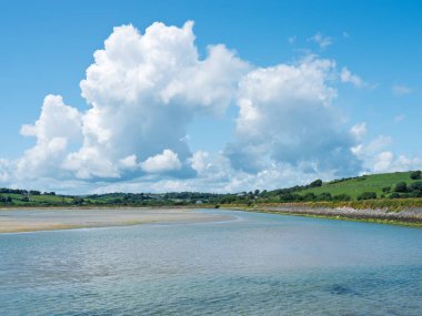 Yumuşak su gelgitlerinin, kumlu bir esnemenin ve parlak, geniş gökyüzünün altındaki yemyeşil tepelerin olduğu canlı bir manzara..