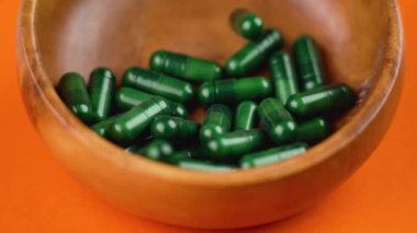Yeşil takviye kapsüller parlak turuncu arka planda ağır çekimde kırsal ahşap bir kaseye düşüyor. Döndürme