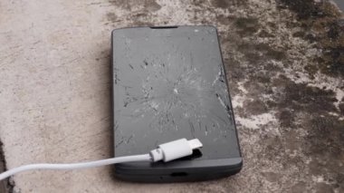 Dış beton grunge yüzeyinde beyaz şarj kablosu olan siyah kırık bir akıllı telefonun kırık cam dokunmatik ekranı. İletişim sorunları kavramı