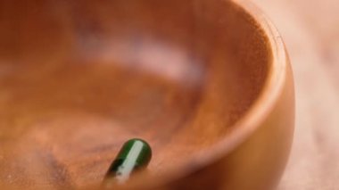 Bitkisel yeşil destek kapsülleri yavaş çekimde ahşap bir kaseye düşüyor. Kapat.