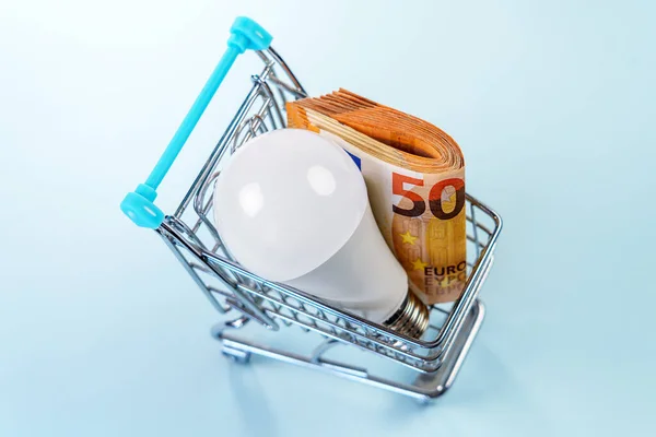 Mini supermarket shopping trolley with electric energy saving light bulb and euro money. Wad of cash banknotes. Concept of financial discount for electricity pay. Cheap reduced tariff idea