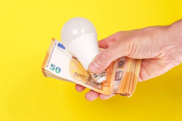 Electric energy saving light bulb with euro bills in hand on a bright yellow creative background. Conceptual consumer payment idea