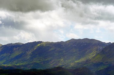 Yokuşlarda bitki örtüsü olan bulutlu bir gökyüzündeki dağ zirvelerinin dramatik manzarası