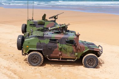 Gijon, İspanya - 24 Mayıs 2024: İki askeri zırhlı araç arka planda okyanus olan kumlu bir plaja park edildiler. Makinelerin ikisi de kamufle edilmiş ve üzerine silah monte edilmiş.