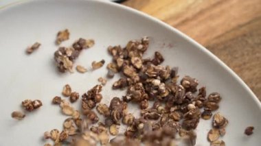 Kuru organik granola bir tabağa dökülüyor. Kaba doku, çeşitli taneler ve çikolata parçaları. Besleyici ve enerji dolu bir kahvaltı için mükemmel. Ağır çekim. Üst görünüm