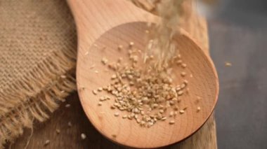 Organik susam tohumlarının yakını kırsal bir masaya yerleştirilmiş tahta bir kaşığa dökülüyor. Çuval bezi arka plana doku ekler. Doğal gıda konsepti. Ağır çekim. Döndürme