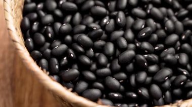 Tahta yüzeyde dokuma sepetinde çiğ siyah fasulye. Koyu renkli doğal baklagiller. Organik malzemeler. Üst Manzara. Döndürme
