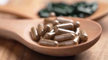 Kahverengi bitkisel kapsüller tahta bir kaşığa düşüyor. Arka plandaki küçük tepside yeşil süper yiyecek hapları var. Makro atış. Yavaş çekim