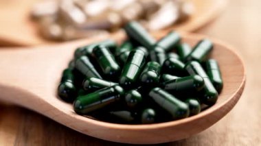 Koyu yeşil kapsüller, ahşap bir kaşıkta, kırsal bir yüzeyde. Arka planda bitkisel haplar olan bir kap daha. Doğal destek konseptinin çeşitliliği. Makro atış. Döndürme