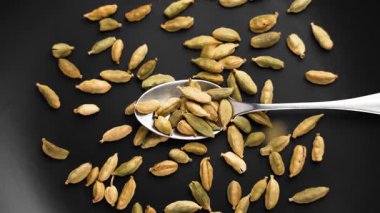 Koyu seramik yüzeyine düşmüş yeşil kakule kapsülleri olan metal kaşık. Dağınık aromatik baharat. Yakın plan çekim. Yavaş çekim
