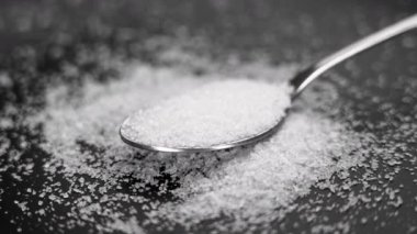 Kristalize edilmiş tuz tozu kaşıkta ve siyah seramik yüzeye saçılmış. Döndürme. Makro