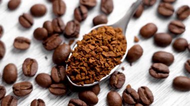 Kavrulmuş kahve çekirdekleri ve beyaz tahtanın üzerinde metal çay kaşığı içinde kahverengi çözünebilir granüller. Döndürme