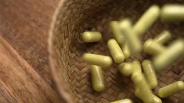 Sarı yeşil ilaç kapsülleri ahşap masada ağır çekimde hasır bir kaseye düşüyor. Üst görünüm