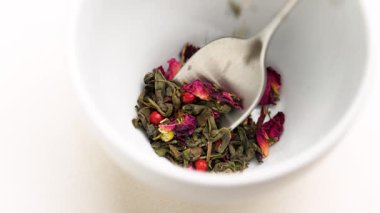 Düşen çiçek yaprakları ve yeşil çay yaprakları beyaz seramik fincanda aromatik şifalı içecek hazırlamak için çay kaşığı ağır çekimde.
