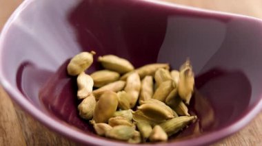 Kurumuş organik kakule kapsülleri ağır çekimde ahşap masada kırmızı seramik kaseye düşüyor.