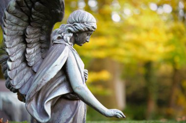 Bir sonbahar mezarlığındaki kanatlı bir melek figürü uzanmış elleriyle üzgün bir şekilde yere bakar.