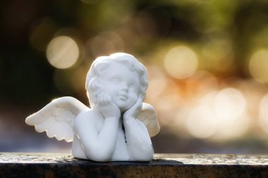 Mezar taşındaki küçük beyaz bir melek figürü Bokeh baloncukları olan bulanık bir arka plana karşı gökyüzüne doğru uzun uzun bakar.