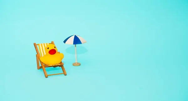 Plaj rahatlaması ve tatil konsepti. Oyuncak güneş şezlongu, ördek ve şemsiye mavi renkli kağıt arka planda.