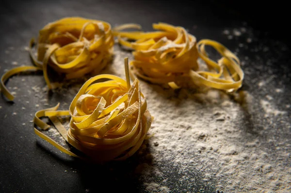 Taze pişmiş makarna, un tozuyla kaplanmış karanlık bir yüzeyde yatıyor. İtalyan makarnası. Tagliatelle. Çiğ makarna. İtalyan makarna tarifi.