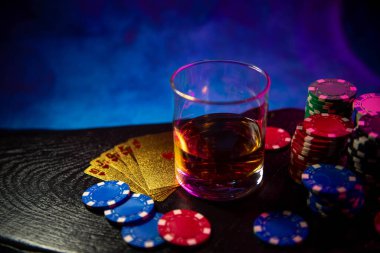 Oyun çipleri masanın üzerinde, siyah tahta bir yüzey üzerinde bir bardak alkolün yanında yatıyor. Kumar oynamak için oyun fişleri. Poker fişleri. Kartlar oynanıyor.